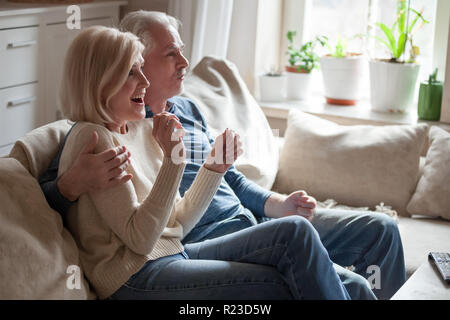 Excited aged couple happy watch romantic movie relaxing on couch, smiling elderly husband and wife supporting sports team resting on sofa spending wee Stock Photo