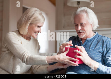 Smiling elderly wife making birthday surprise for beloved husband, aged man get present from loving spouse on special occasion, caring senior couple e Stock Photo