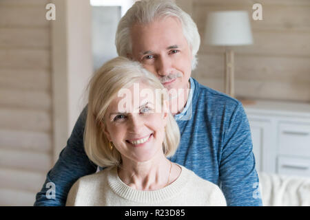 Portrait of happy aged couple hugging posing for family picture at home, caring senior husband embrace wife from behind shooting together, loving elde Stock Photo