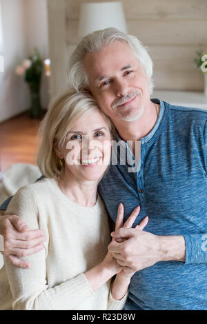 Portrait of smiling aged husband and wife embracing making family picture at home, happy senior couple hugging and holding hands looking at camera sho Stock Photo