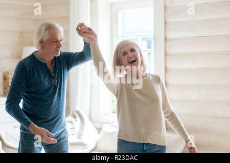 Happy senior husband and wife have fun spending time in country house together, smiling aged couple dance swirling and swaying at home, excited elderl Stock Photo