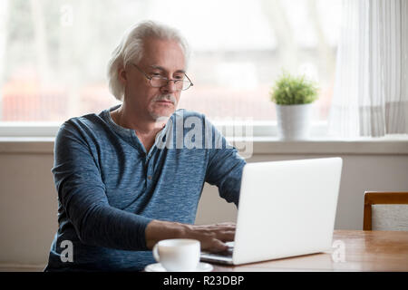 Serious aged man busy working at laptop drinking morning coffee at home, thoughtful senior male use computer writing email message, typing on pc or br Stock Photo