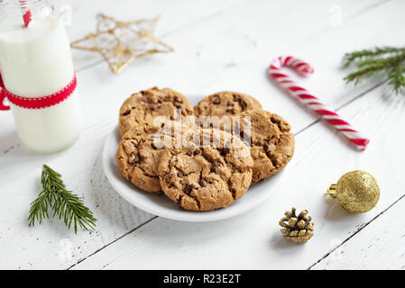 Chocolate chip cookies and Milk for Santa. Traditional Christmas homemade cookies, candy cane and bottle of milk with Christmas decor on white wooden  Stock Photo