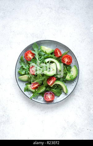 Green salad with sliced avocado, tomatoes and fresh arugula on white stone table with copy space. Healthy diet vegan vegetarian summer vegetable salad Stock Photo