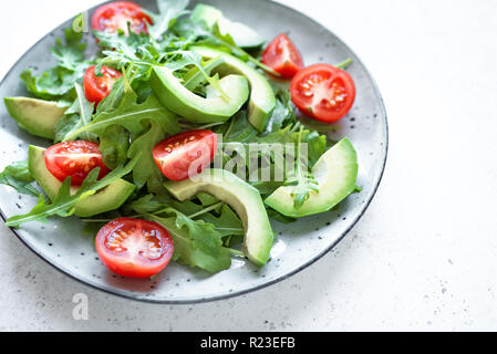 Green salad with sliced avocado, tomatoes and fresh arugula on white stone table, close up. Healthy diet vegan vegetarian summer vegetable salad food  Stock Photo