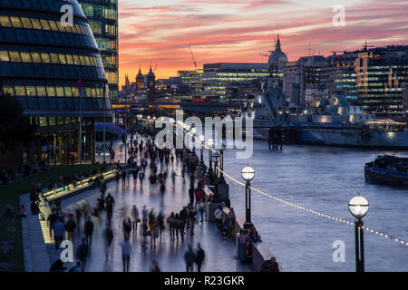 London, England, UK - September 27, 2018: Pedestrians and tourists walk along the River Thames Path beside City Hall and HMS Belfast at sunset. Stock Photo