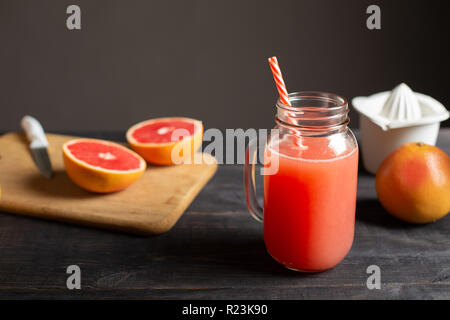 Freshly squeezed grapefruit juice in a jar with a handle. On a black wooden table are whole and sliced grapefruits and white manual juicer. Stock Photo