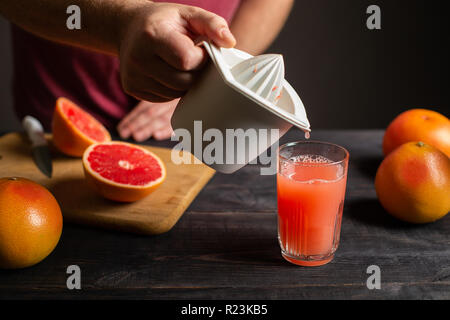 Freshly squeezed grapefruit juice is poured from a manual juicer in a glass. White plastic squeezer in male hand. On a black wooden table are whole an Stock Photo