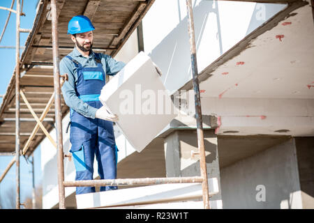 Builder warming a building facade with foam panels standing on the scaffoldings on the construction site Stock Photo