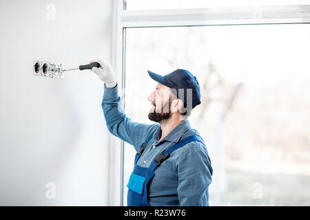 Electrician in uniform mounting electric sockets on the white wall indoors Stock Photo