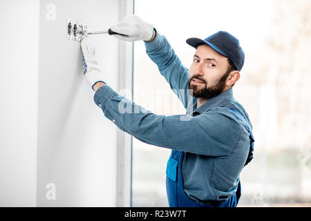 Electrician in uniform mounting electric sockets on the white wall indoors Stock Photo
