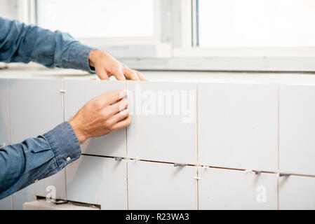 Workman in blue uniform inserting distance crosses installing ceramic tiles indoors, close-up view focused on the hands Stock Photo