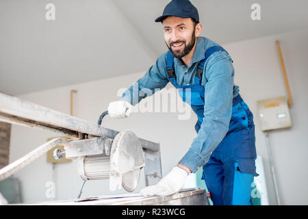 Builder in uniform cutting tiles with big electro saw on the construction site outdoors Stock Photo