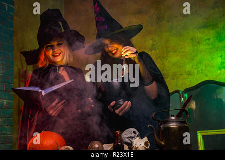 Portrait of smiling two witches in black hats reading book at table with pumpkin and skulls in dark room Stock Photo