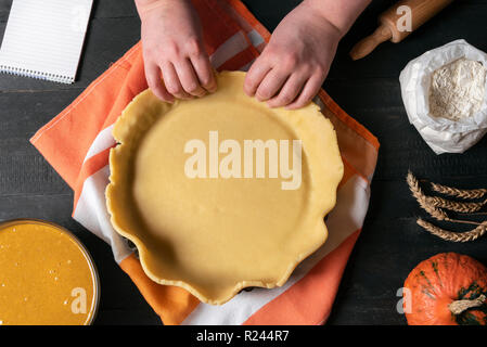 Woman hands working a pie dough in a tray, on a kitchen table, surrounded by pumpkin pie ingredients and a notebook. Traditional pies baking. Stock Photo