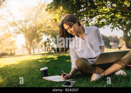businesswoman sitting on grass at park with laptop and making notes in her diary. Freelancer female working while sitting at park. Stock Photo