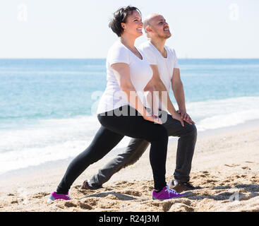 Cheerful smiling mature couple practicing yoga exercises on the sandy beach Stock Photo
