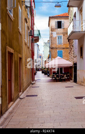 Cozy and empty narrow street between two stone houses with orange doors and shutters on a summer morning. Small cafe and an umbrella in the corner. Za Stock Photo