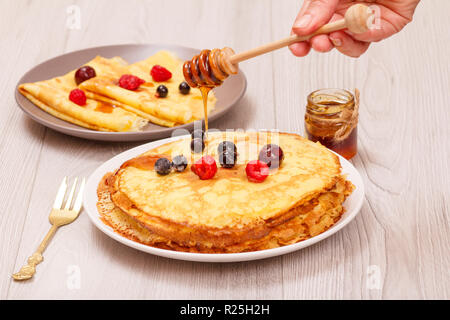 Woman's hand holding a wooden spoon of honey with homemade pancakes topped with frozen raspberries and blackberries on the background. Stock Photo