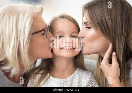 Smiling kid girl enjoys mom and grandma kissing on cheeks Stock Photo