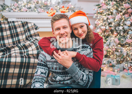 Picture of happy couple posing together on camera. and smiling. She stands behind and embrace him. Man touches her hand. There are fireplace and Chris Stock Photo