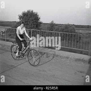 1960s, attractive young woman riding a bicycle on a path across a bridge in the countryside. Stock Photo