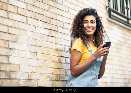 Happy Arab girl using smart phone on brick wall. Smiling woman with curly hairstyle in casual clothes in urban background. Stock Photo