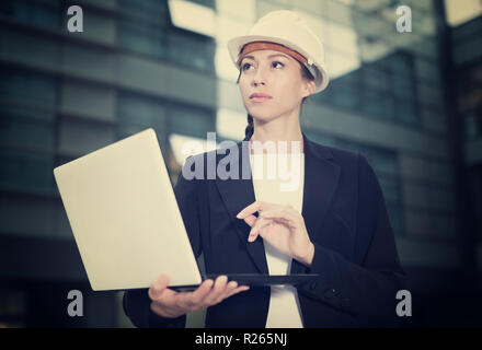 Smiling woman architector in suit and hat is exploring project in her laptop near the building. Stock Photo