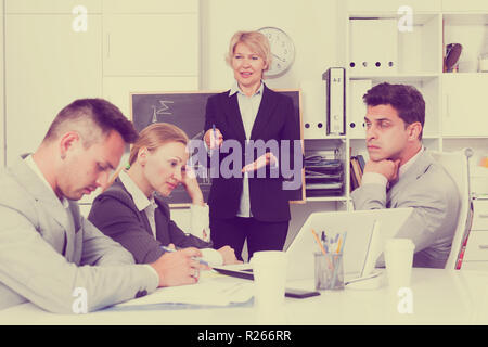 Mature businesswoman standing near blackboard in office, expressing dissatisfaction with work of colleagues Stock Photo