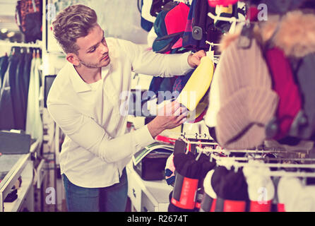 Smiling guy deciding on warm hat in sports store Stock Photo
