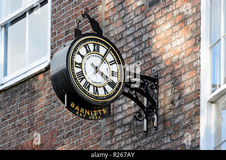 Close-up of public clock telling the time, mounted high on brick wall outside Barnitt's home and garden shop - York, North Yorkshire, England, UK. Stock Photo