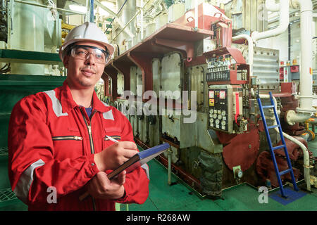 Ship's mechanic near marine diesel generators on a merchant ship in the engine room with all the piping, generators, turbins, etc. writing something d Stock Photo