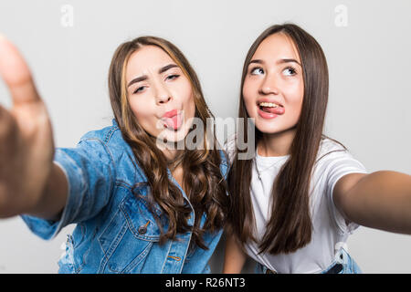 Portrait of two joyful multiracial women with black and blonde hair taking selfie on smartphone, with tongue out showing victory sign isolated over bl Stock Photo