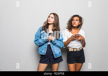 Two young best friends women are busy at their mobile phones in social nets, wearing casual shirts, isolated on white background Stock Photo