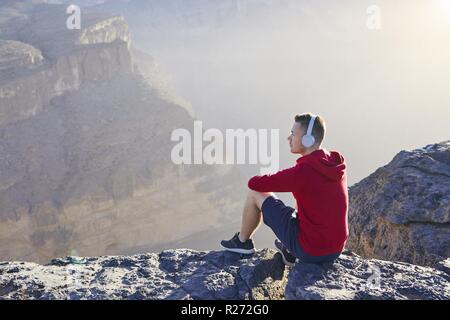 Relaxation in mountains. Young man with headphones sitting on the edge of cliff and listening music. Stock Photo