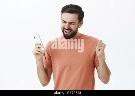 Waist-up shot of enthusiastic pleased and delighted carefree charming adult male in wireless earphones holding smartphone dancing in rhythm of music s Stock Photo