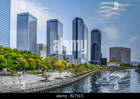 Modern buildings of a commercial area next to a river lined with green trees a boat navigates the river, beautiful day of blue sky. In Osaka, Japan Stock Photo