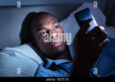 African Man Lying On Bed Using Mobile Phone At Night Stock Photo