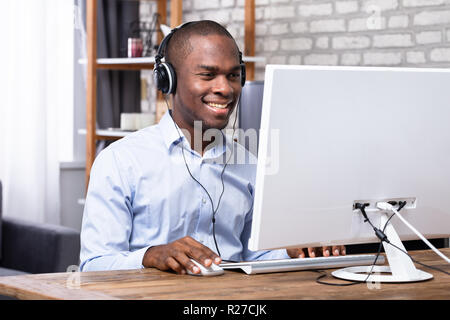 Smiling Man Listening To Music On Headphone While Using Computer Over Wooden Desk Stock Photo