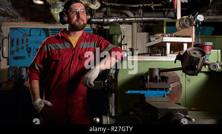 Worker in red uniform operating in manual lathe in metal big workshop with dark environment. Stock Photo