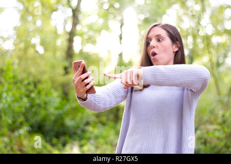 Young caucasian european brunette girl pointing her cellphone very surprised in the forest or nature Stock Photo