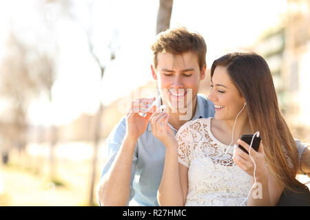 Happy couple sharing earbuds to listen to music from a smart phone sitting on a bench in a park Stock Photo