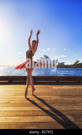 Young fit ballerina in red tutu dancing on timber deck at the shores of Sydney Harbour in soft morning light under blue sky. Stock Photo