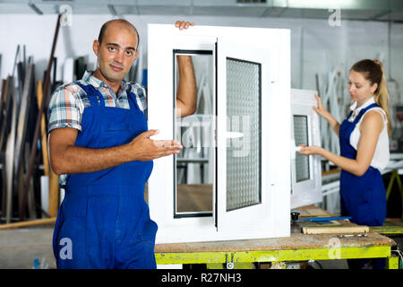Happy workman in blue overalls demonstrating pvc window in workshop Stock Photo