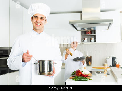 Male cook wearing uniform with stewpot working on kitchen with woman cook Stock Photo
