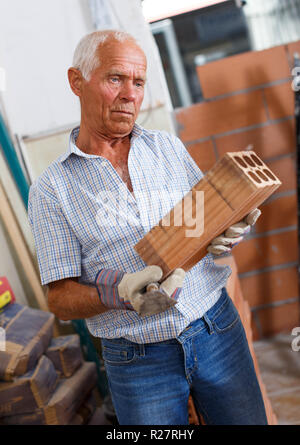 Skillful older man inspecting brick for installing wall in building under construction Stock Photo