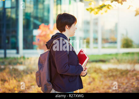 Lifestyle outdoors portrait of young student holding books and backpack. Autumn side view of thoughtful woman in university campus. Stock Photo
