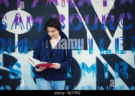 Casual woman student reading book, outdoors lifestyle portrait leaning back on graffiti wall background.