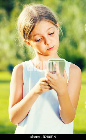 girl in elementary school age is very surprised while looking at mobile phone on summer day Stock Photo