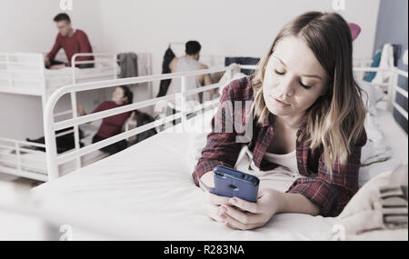 Attractive girl with phone in hands lying on top bunk of bunk bed in bedroom of hostel Stock Photo